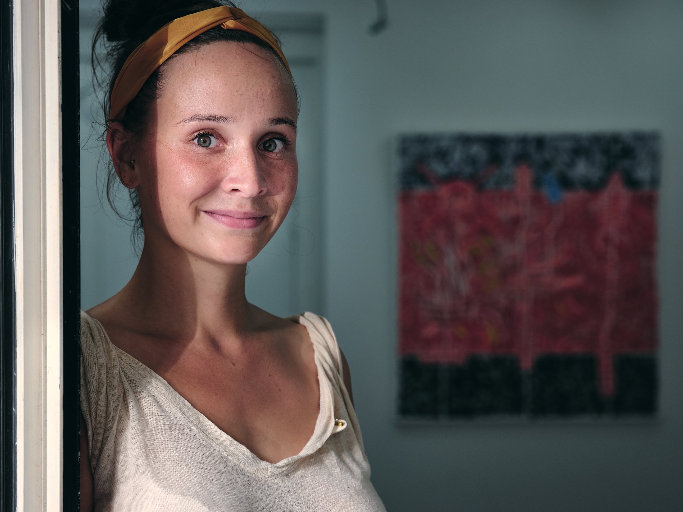 Portrait — femme au bandeau jaune sur un seuil, œuvre accrochée au mur derrière elle — photographie Romain Cruse
